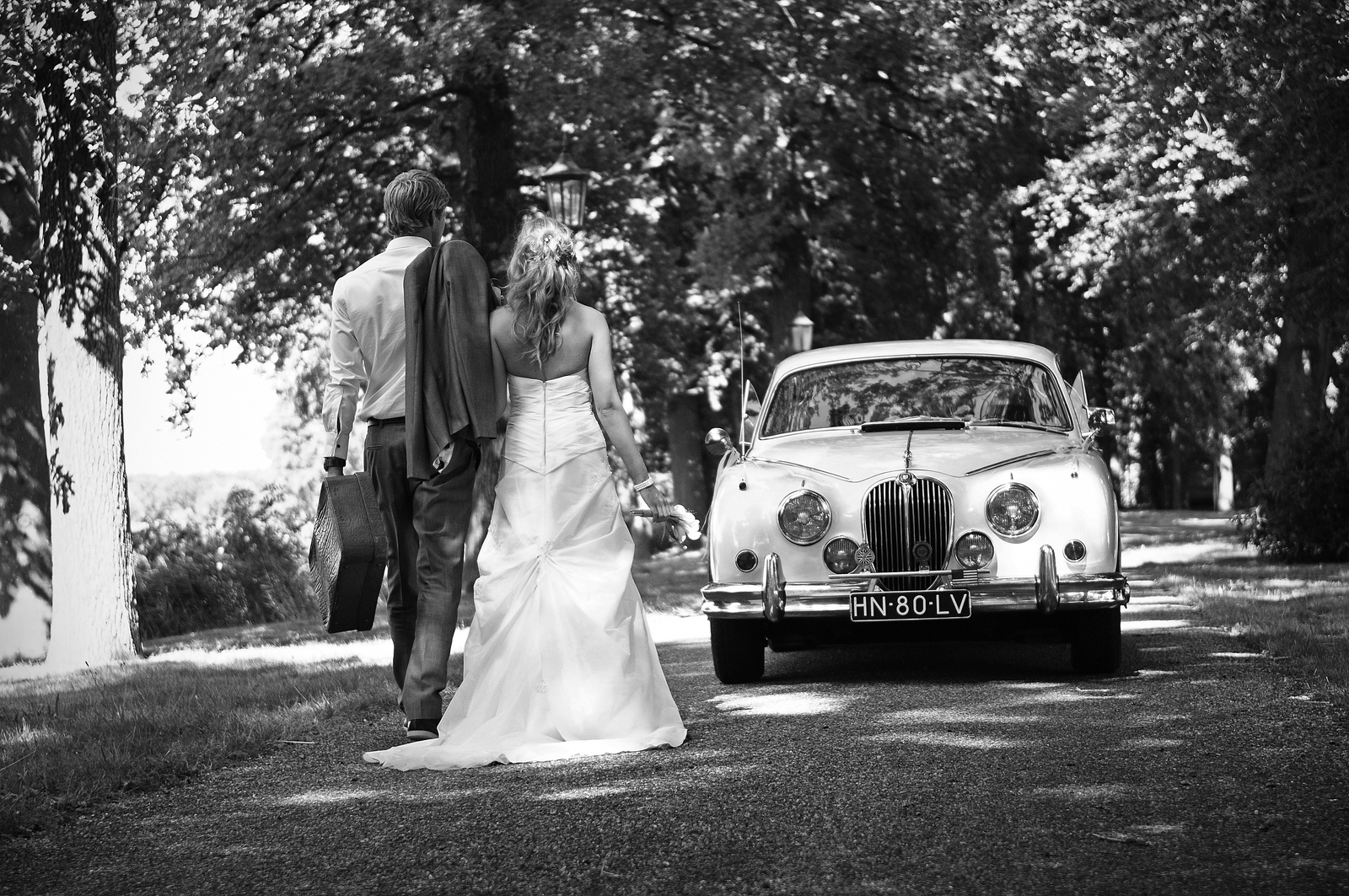 Bruidspaar loopt met koffer naar hun witte Jaguar MK2 2.4 trouwauto uit 1961